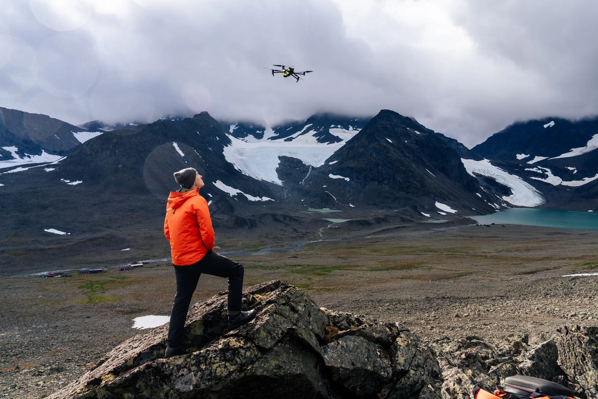 Erik Schytt Mannerfelt i orange jacka på en klippa, tittar på en drönare som flyger över glaciärerna och en smältvattensjö
