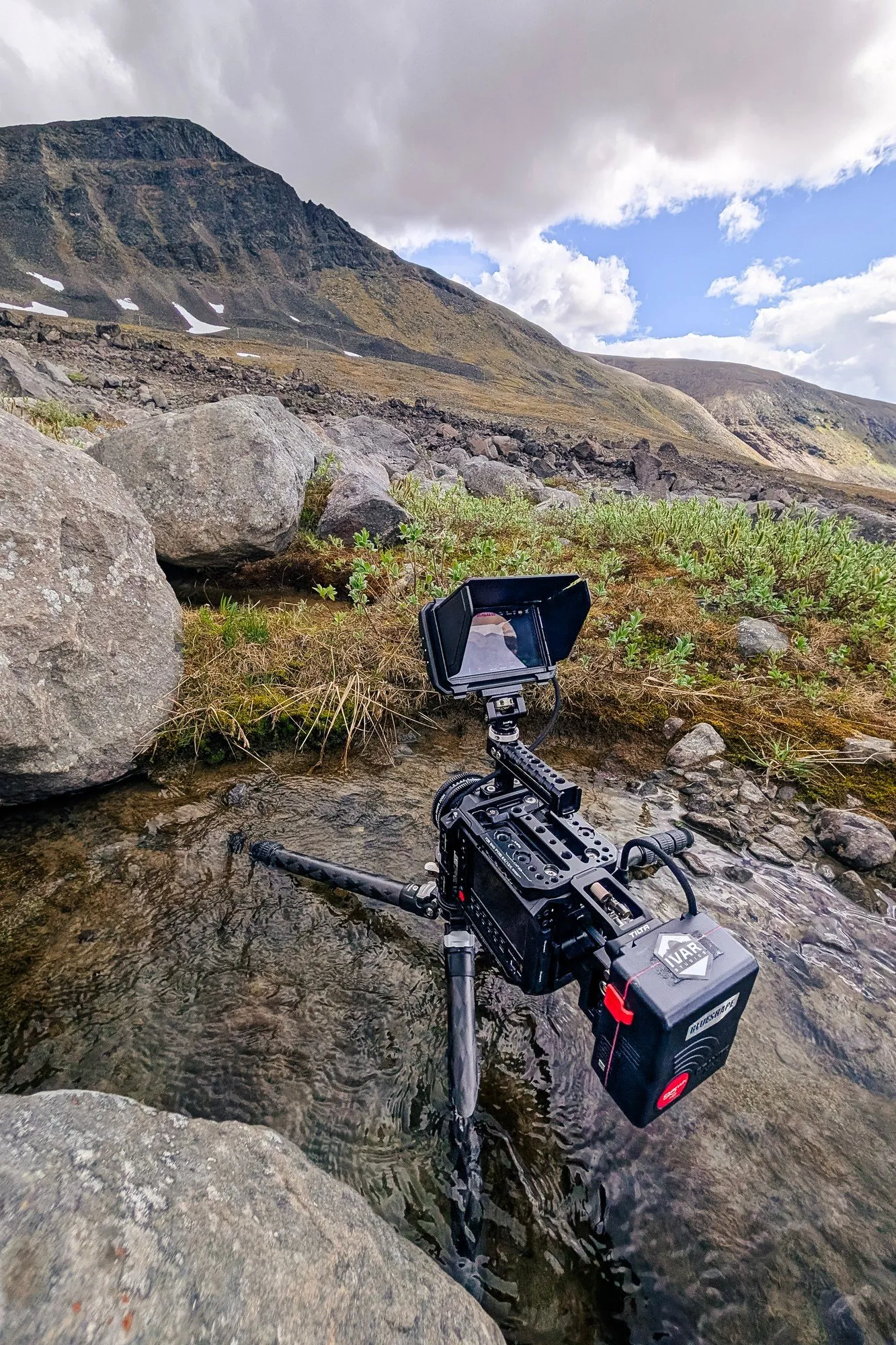 En filmkamera med skärm på stativ står i en grund fjällbäck bland stenblock