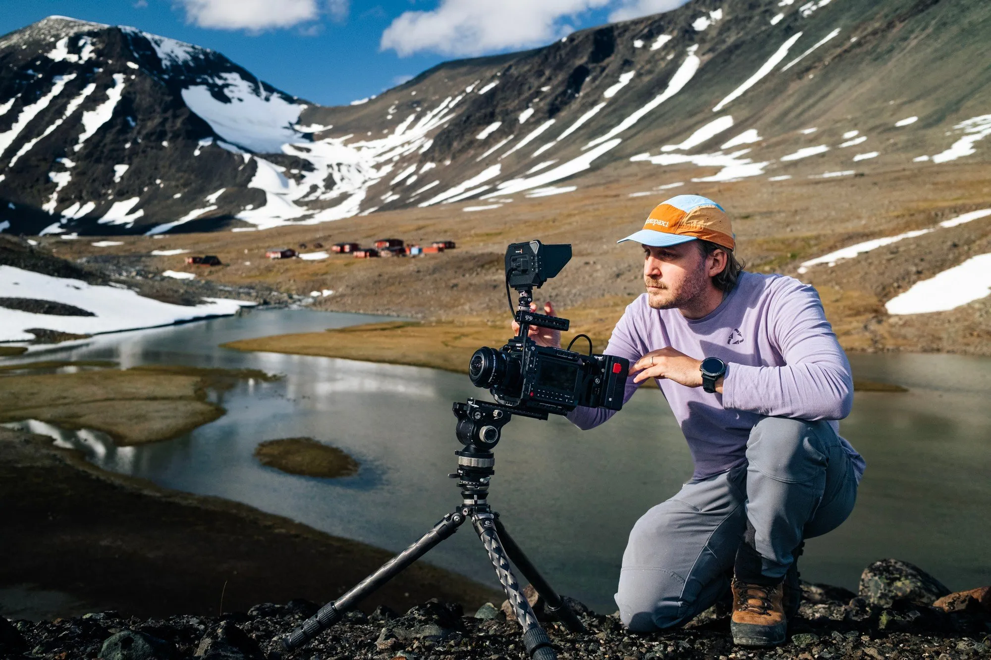 En filmare i keps knäböjer vid en filmkamera på stativ vid en stilla glaciärtjärn, med röda stationshus och snöiga berg bakom
