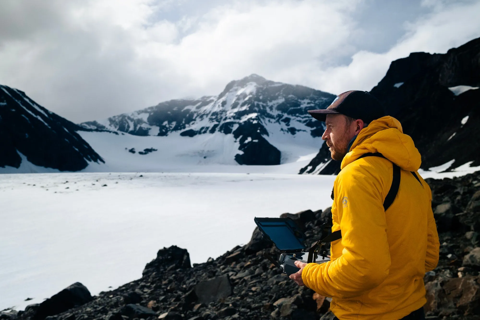 Martin Edström i gul jacka håller en drönarkontroll på stenarna vid Storglaciären, med glaciären och snöiga toppar bakom sig