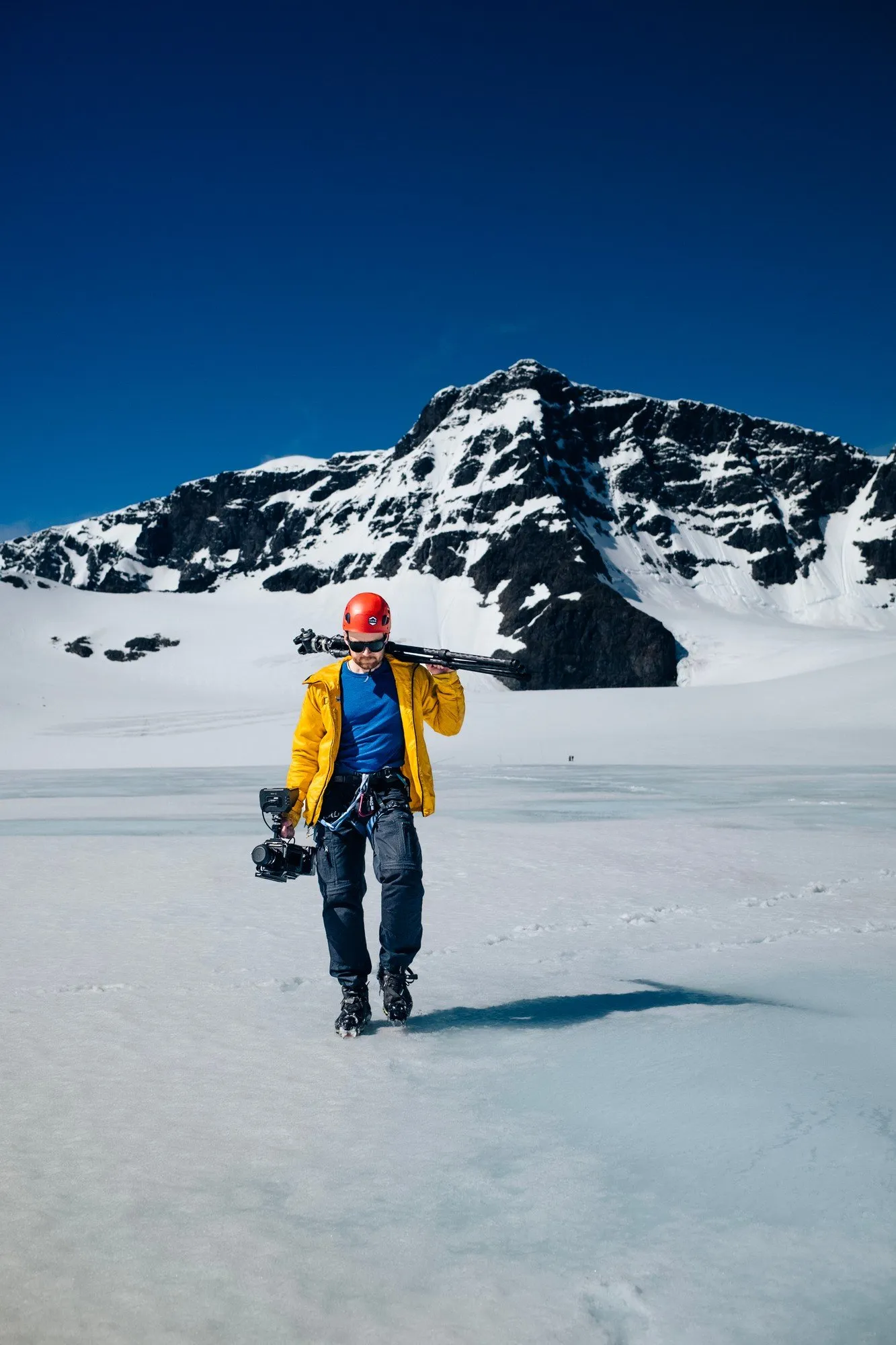En person i hjälm bär kamerautrustning över Storglaciären under blå himmel